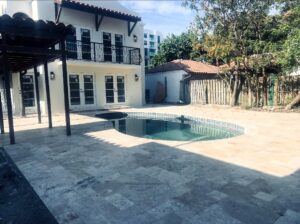 Kidney-shaped pool in a paved courtyard beside a two-story building with glass doors and a covered patio area on the left, outdoors.