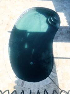 Kidney-shaped swimming pool with dark water, tiled coping, and a light stone patio; shadows fall across the pool surface.