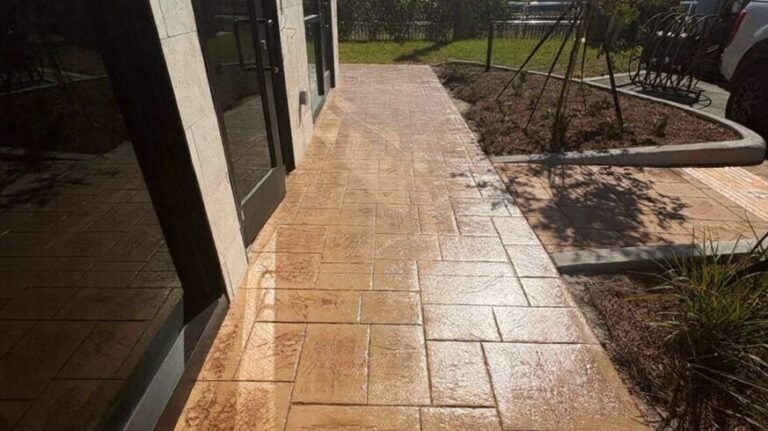 Sunlit tan stamped concrete walkway along a glass storefront with a small garden bed on the right and a car in the background.