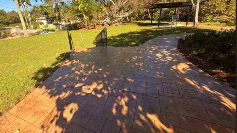 Sunlit park walkway with a curved tiled path, dappled tree shadows, and a grassy area on the left; a railing leads toward the lawn.