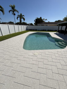 A backyard swimming pool surrounded by newly installed light gray interlocking stone pavers, completed by Cobra Construction in Delray Beach, Florida. The pavers are arranged in a clean herringbone pattern and meet the curved edge of the pool. A white vinyl privacy fence encloses the manicured green lawn in the background under a clear blue sky with palm trees.