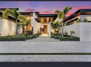 The modern, luxury front entrance of a home featuring large light gray driveway pavers and a matching stone walkway, constructed by Cobra Construction in Delray Beach, Florida. The contemporary estate showcases crisp white walls, bold black trim, and natural warm wood ceilings above the entryway. Lush tropical landscaping, including vibrant green hedges and tall palm trees, frames the entrance against a dramatic pink and purple sunset sky.