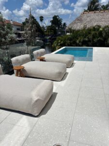 A modern poolside patio featuring large, square, off-white shell stone pavers expertly installed by Cobra Construction in Delray Beach. The pavers showcase a distinct natural texture embedded with fine coquina and fossilized shell fragments. Placed on the stone deck are three plush, neutral-toned outdoor lounge daybeds next to a vibrant blue swimming pool, highlighting a luxury backyard hardscape design.