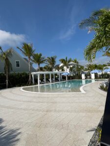 A large, luxury community swimming pool surrounded by a custom paver deck expertly installed by Cobra Construction in Delray Beach. The hardscape showcases light grey, textured stone pavers laid in a precise herringbone pattern, bordered by concentric, sweeping curved lines that match the pool's zero-entry edge. Palm trees, a white pergola with lounge chairs, and blue umbrellas complete the tropical resort-style landscape.